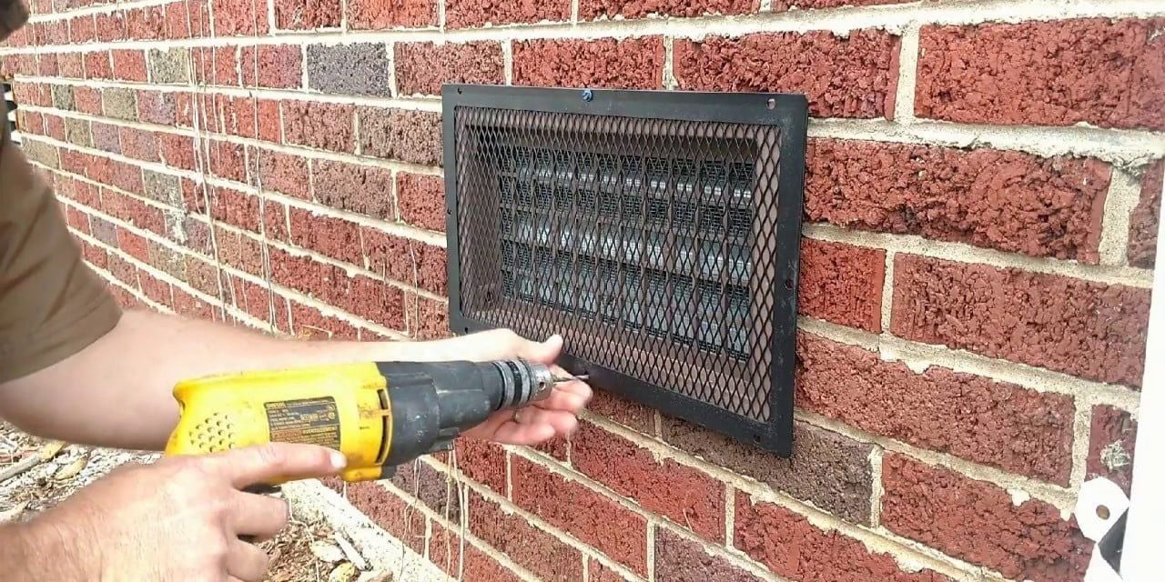 A man using a yellow drill to install a black foundation vent cover over a foundation vent on the side of a brick house.