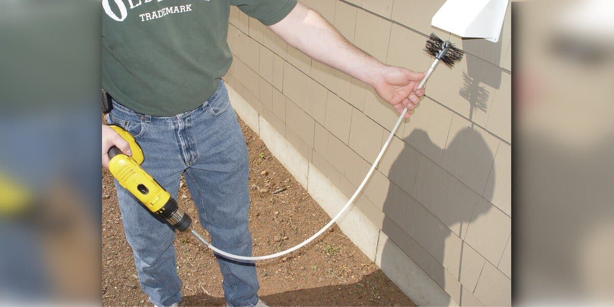 A man holding a dryer vent cleaning kit attached to a yellow drill. He's inserting the auger brush head of the kit into the termination end of the dryer vent.