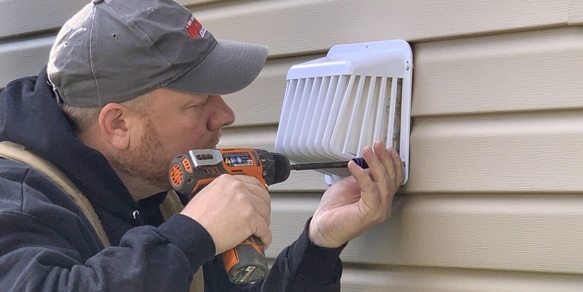 A man in a gray hat installing a code-compliant plastic dryer vent guard onto a house with beige siding using an orange drill.