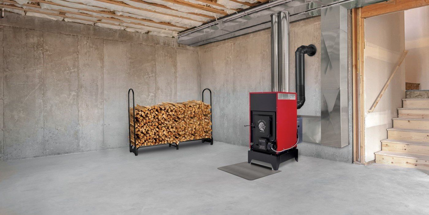 A Fire Chief FC1000E wood burning furnace installed in a basement. There is a rack loaded with firewood not far from the furnace.